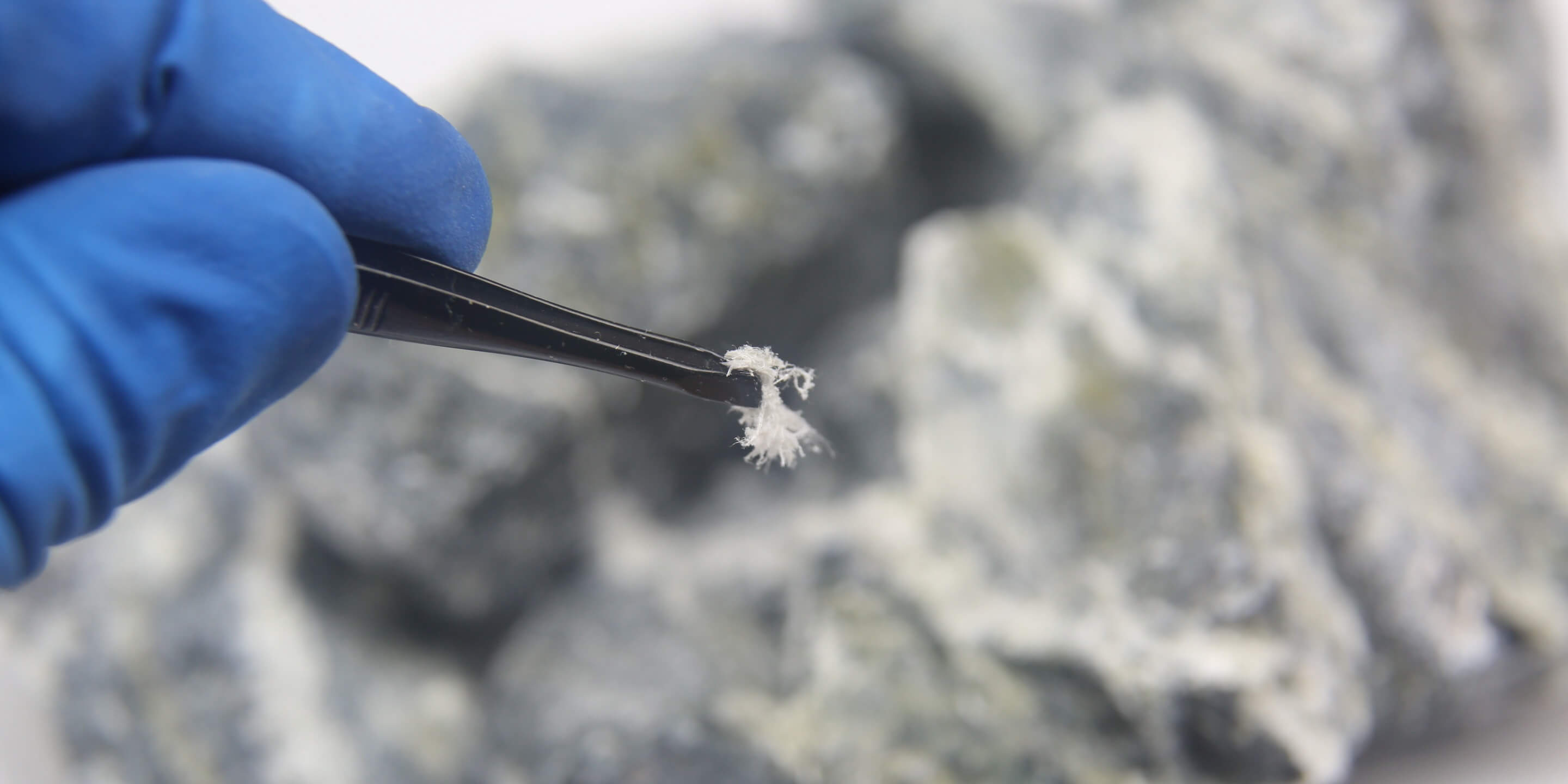 An image of a gloved hand holding up a piece of asbestos with a tweezers.