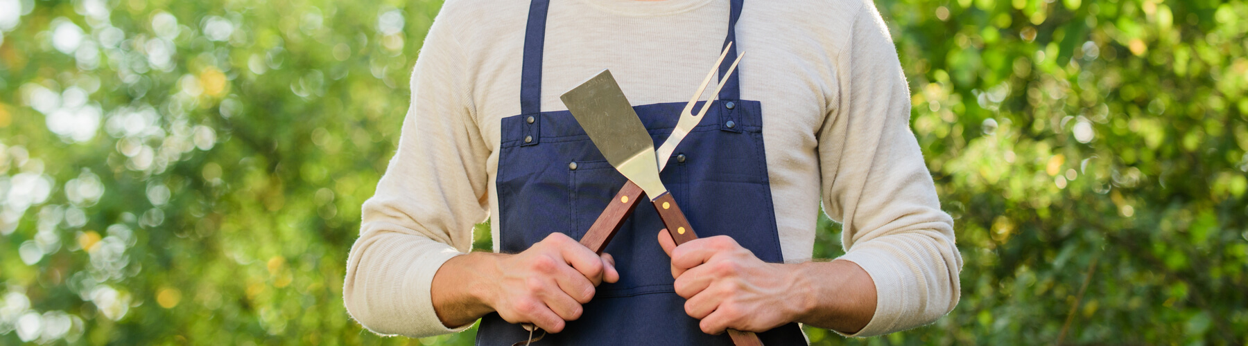 cook off concept, man wearing grilling or cooking apron holding utensils in a cross across chest, challenge