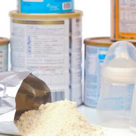 spilled baby formula with backdrop of canisters; hand holding a full scoop next to baby bottle