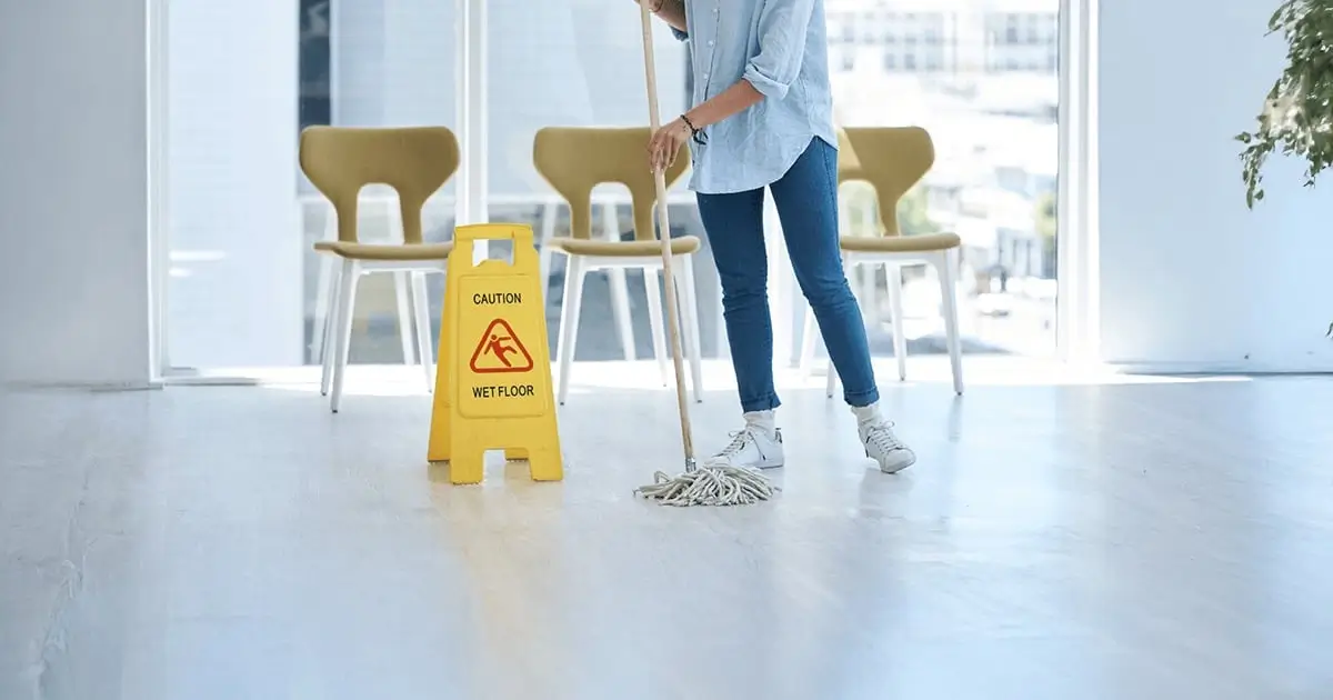Person mopping a wet floor with a caution sign, indicating slip and fall hazard